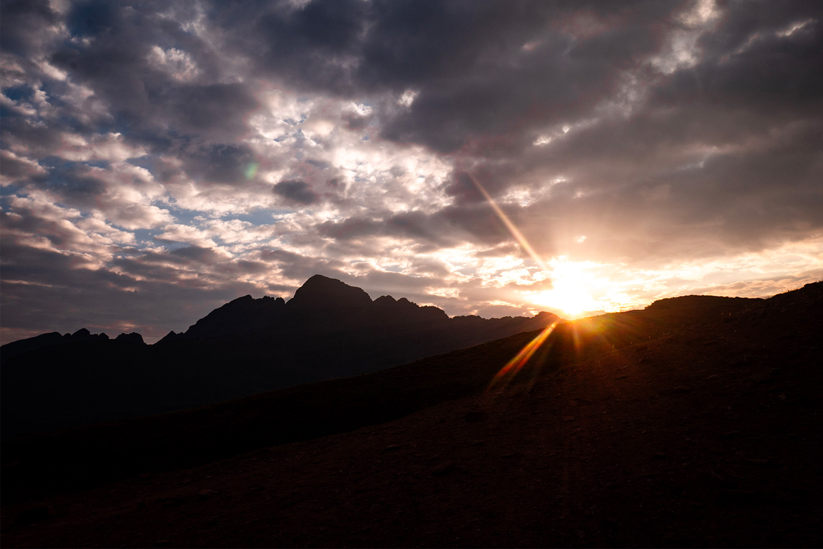 The sun is peaking over the silhouette of ragged mountains. The sky above the sun is cloudy.