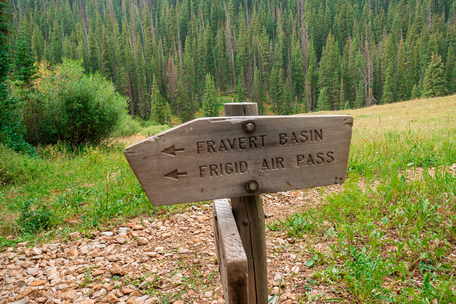 A wooden sign with two arrows pointing to the left read: "Fravert Basin" and "Frigid Air Pass". It is marking a trail junction in the woods.
