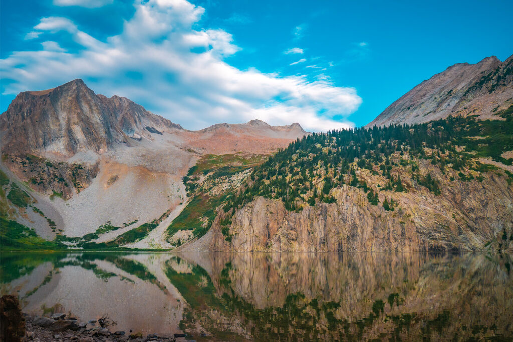 There is a lake in the foreground of the photo with a large cliff and scree fields in the background. The scree fields lead up to several mountain peaks.