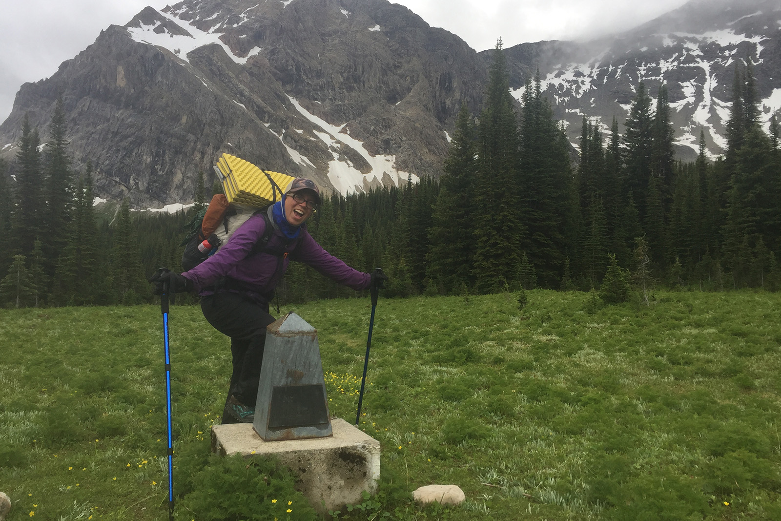 A person wearing a backpack, rain pants, and a rain jacket looking bundled up stands next to a small obelisk. There are pine trees and a mountain in the background and it is a dreary day.