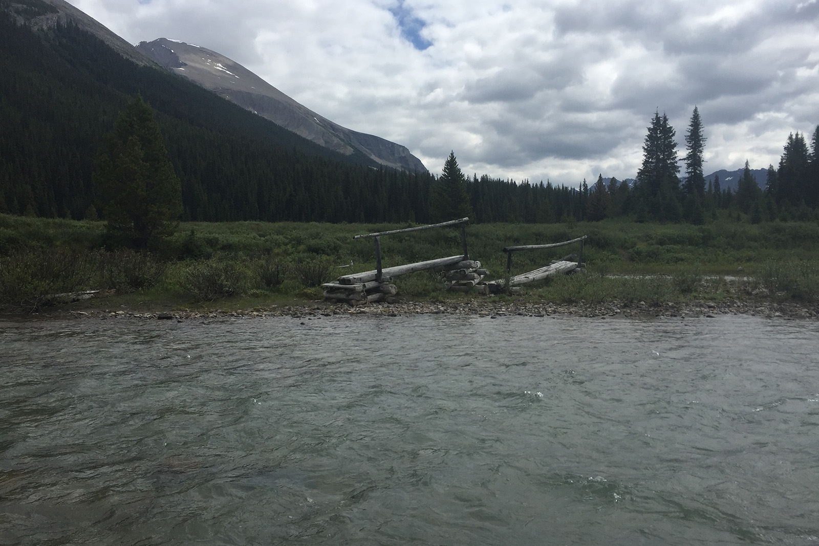 There is a decommissioned bridge on the other side of a fast flowing river. There are trees and mountains in the distance.