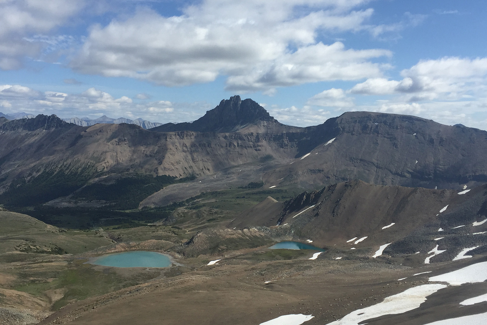 The viewer is standing high up on a ridge looking down towards two blue lakes in the valley below. There are other mountains in the distance.