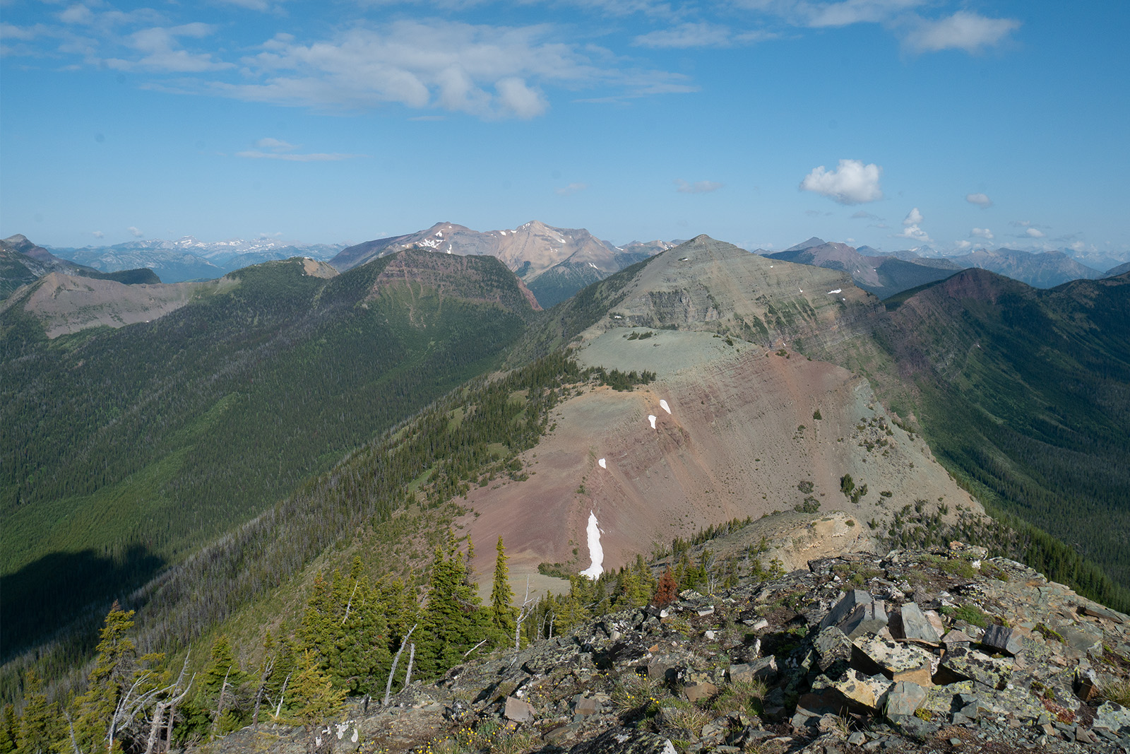 A ridgeline rises and falls steeply between the different peaks. There are other pine covered mountains in the distance.