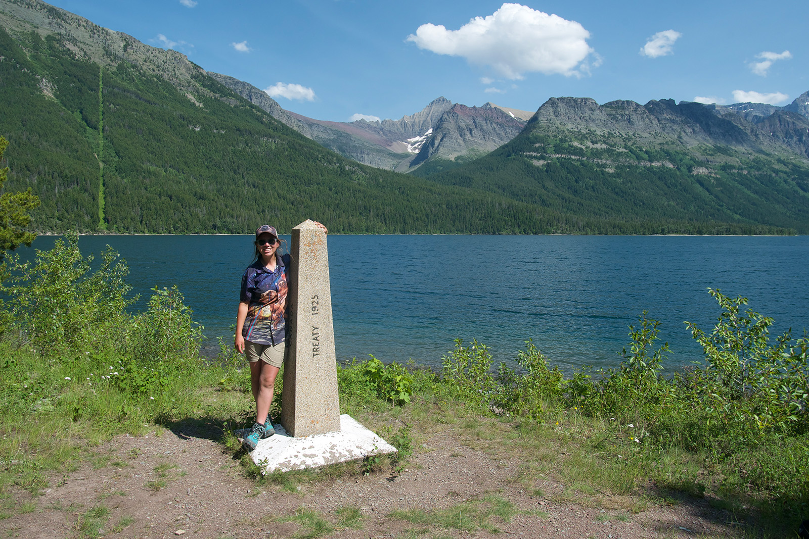 A person stands next to a obelisk that is about the same height. There is a blue lake behind them and pine covered mountains in the distance.