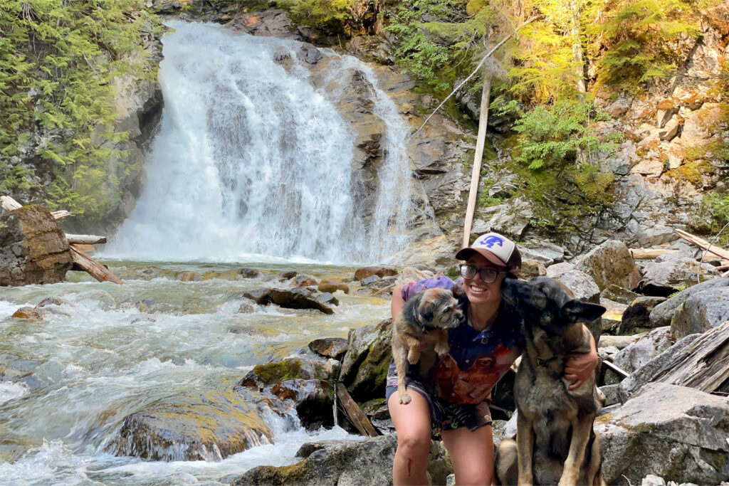 A person is kneeling next to a German Shepherd Dog and holding a Border Terrier in some rocks. Behind them a good sized waterfall drops down a rock face among the trees before flowing past the group.