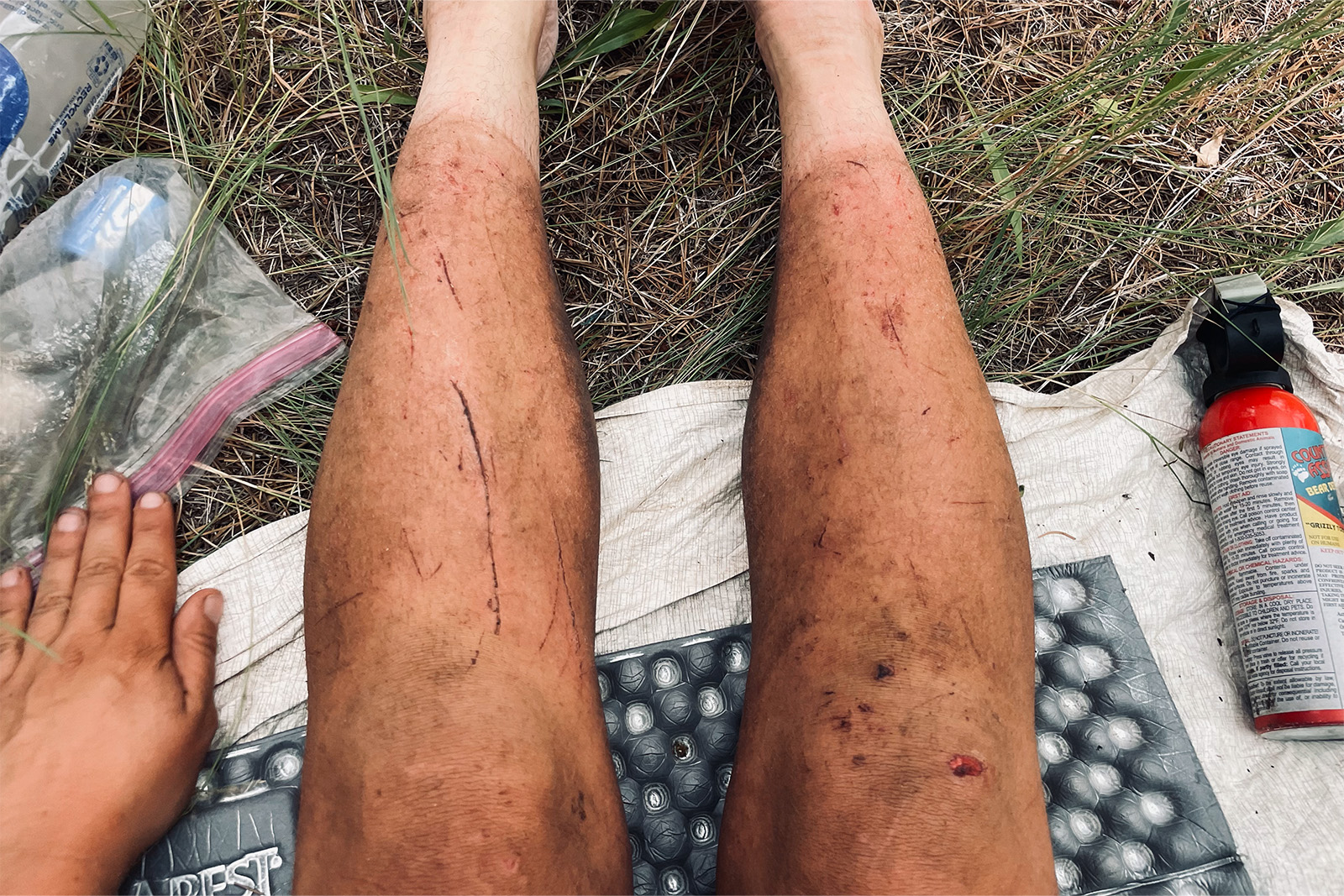 A hikers legs are shown stretched out on a sleeping pad with bear spray on one side and a water filter on the other. They are dirty with many scratches covering them.