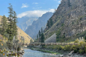 Landscape photo of a wooden suspension bridge spanning a good sized river. Beyond the bridge, granite peaks rise steeply making the valley deep cut.