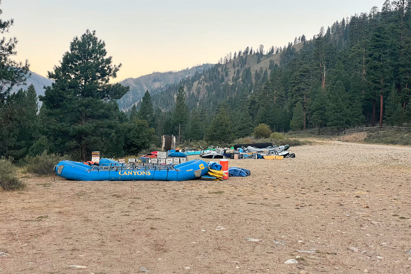 There is an open area of gravel in a pine forest. There are large, blue rafts and lots of gear laid out in the open area.