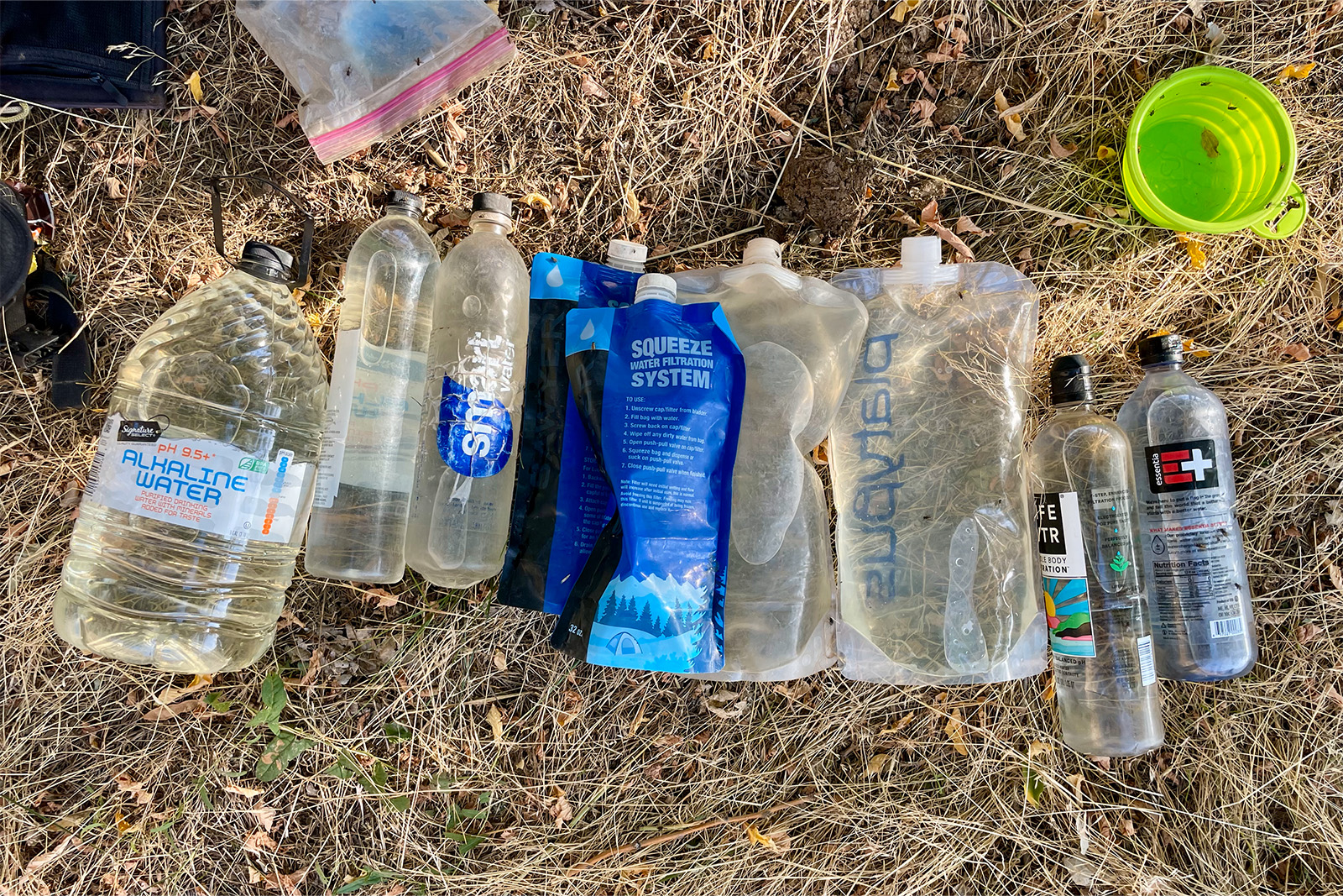 A gallon jug, several smart water bottles, and several water bladders are lined up on the ground surrounded by other pieces of gear. In total, the containers hold about 15 liters of water.