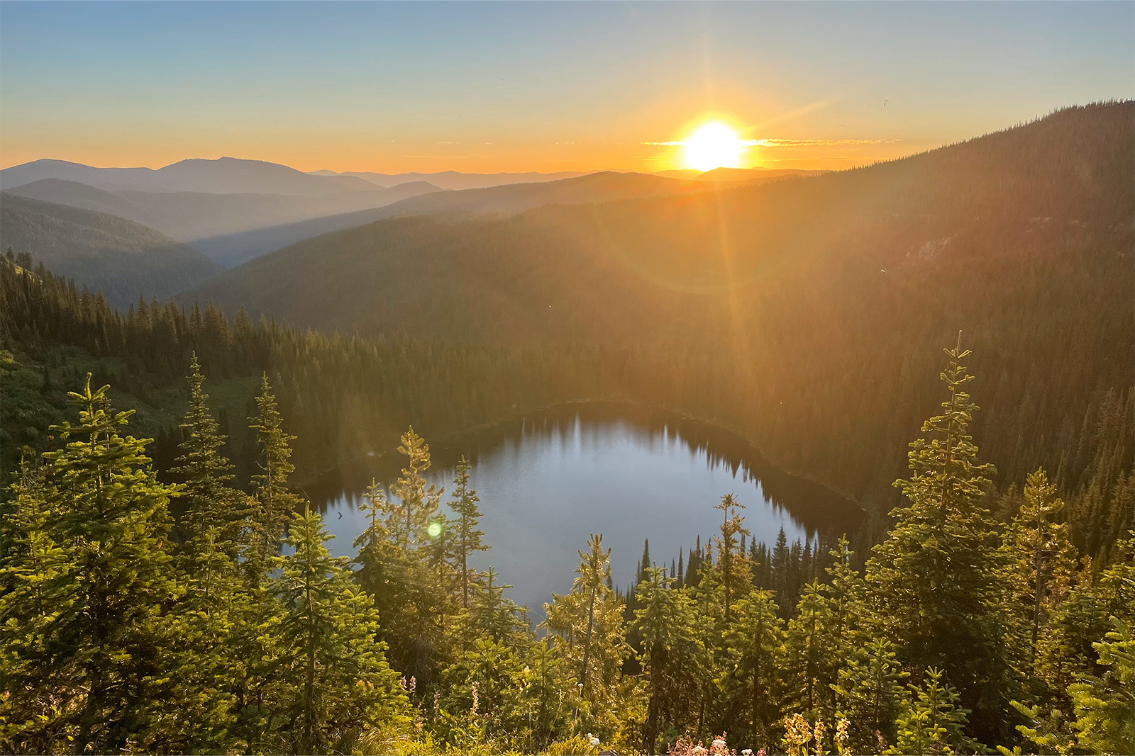 The sun is rising over pine covered ridges to highlight a lake below the viewer. It is a clear day.