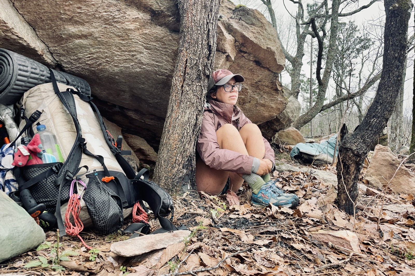 A person wearing a rain jacket huddles next to a rock and their backpack while leaning against a tree. The day is gloomy and the trees are bare.