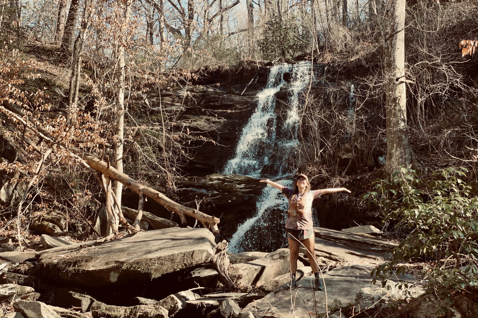 A person is standing below a waterfall looking at the camera with their arms stretched out behind them. There are many large rocks, bare trees, and evergreen bushes in the area.
