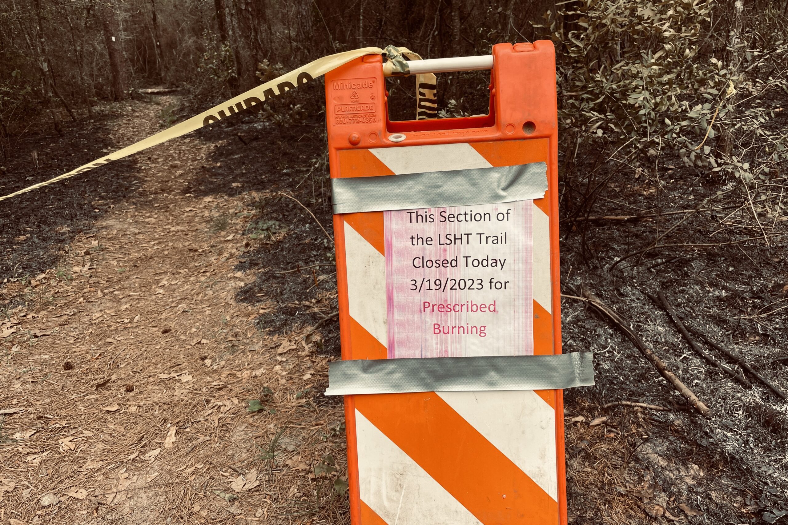 An orange and white sign with caution tape sits in front of the trail. A poster is taped to it and says "This section of the LSHT Trail closed today 3/19/2023 for prescribed burning".
