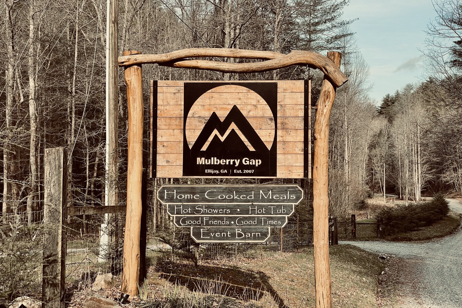 A large, wooden sign for Mulberry Gap Adventure Camp. The camps logo is large and takes up most of the sign. Below it proclaims "home cooked meals, hot showers, hot tub, good friends, good times, event barn". There is a dirt road and forest in the background.