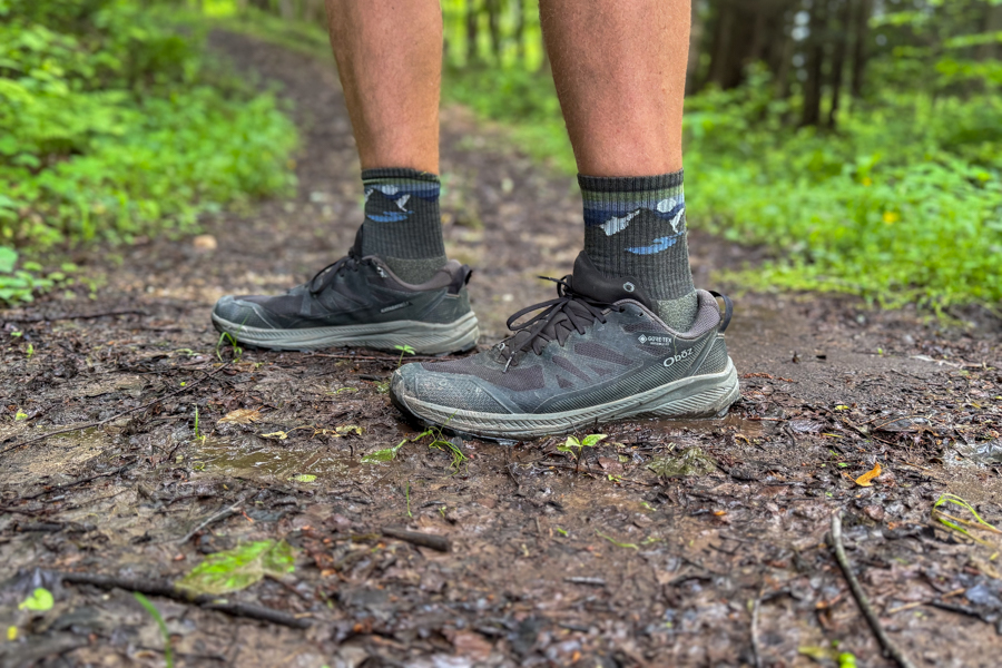 Side view of Oboz Katabatic shoes stepping on muddy forest floor near a small stream.