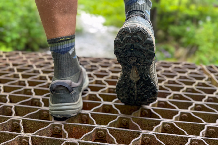 Hiker walking on mixed terrain—mud, rocks, and greenery—wearing Oboz Katabatic shoes.