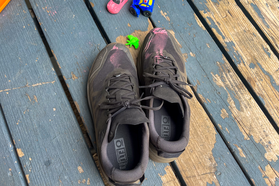 A pair of shoes sits on a deck. The shoes have been colored with red chalk. There are toys laying near the pair of shoes.