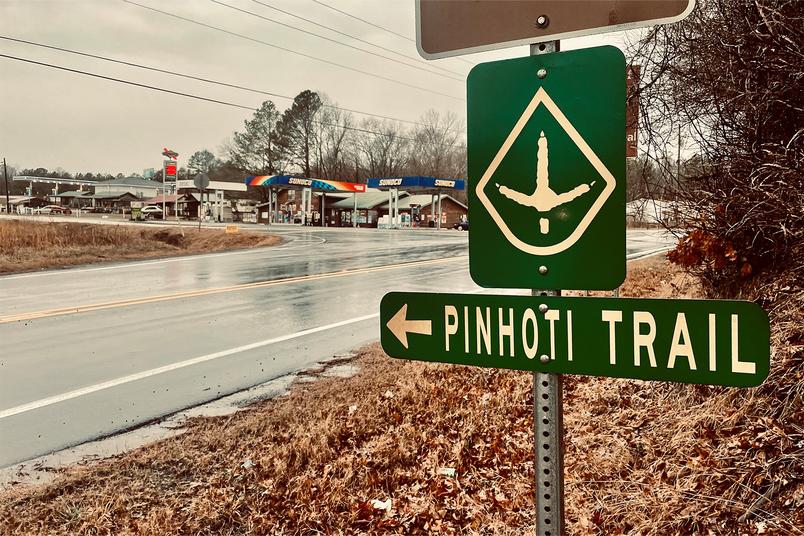 A Pinhoti Trail sign and blaze is bolted to a pole next to a wet road. There is a gas station catty corner from the sign across an intersection. It is raining and the road is wet.