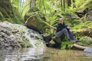 Woman sits next to a creek, cozied up in the Retro Denali Fleece.