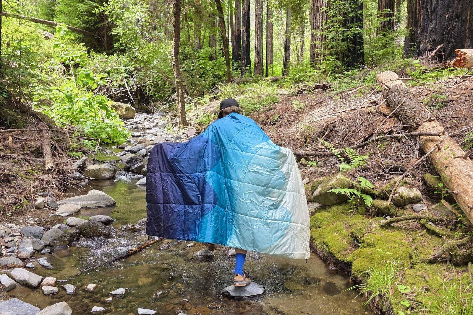 A man walks along a creek while wrapped in a camping blanket.