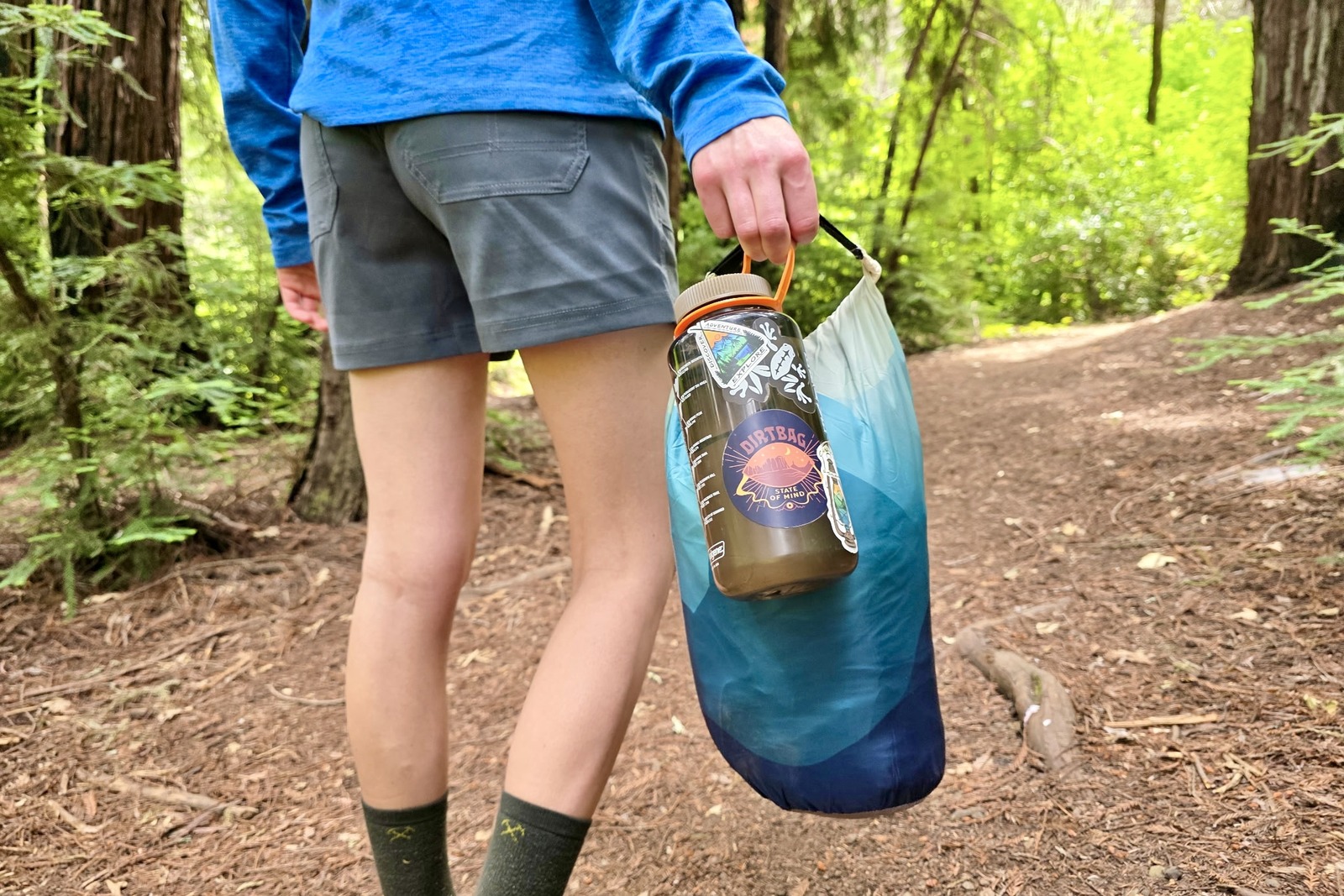 A woman stands holding a blanket in its stuff sack next to a Nalgene to compare size.