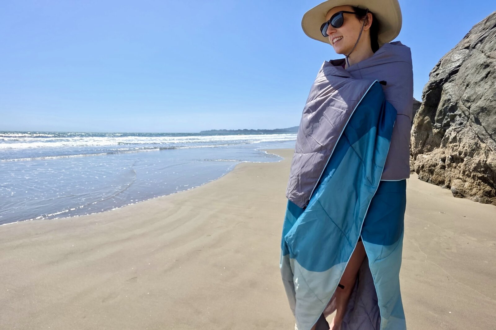 A woman stands wrapped in the Glorsign puffy blanket at the beach.