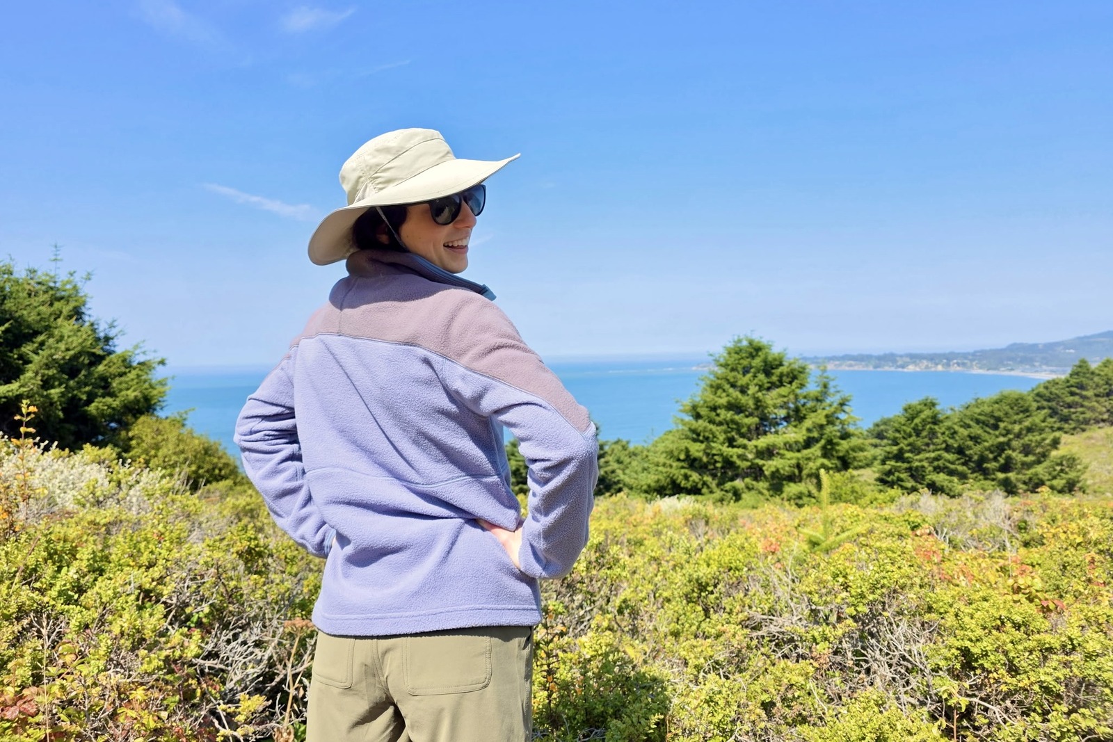 Woman stands over a view of the ocean, showing the back of her fleece sweater.