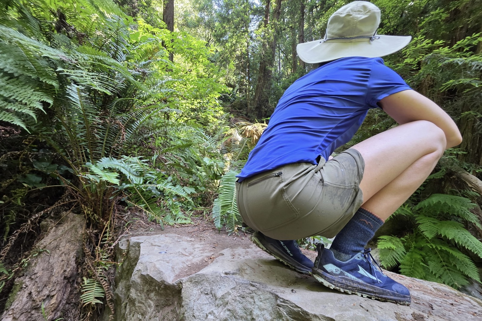 A woman squats on a boulder facing a lush forest.