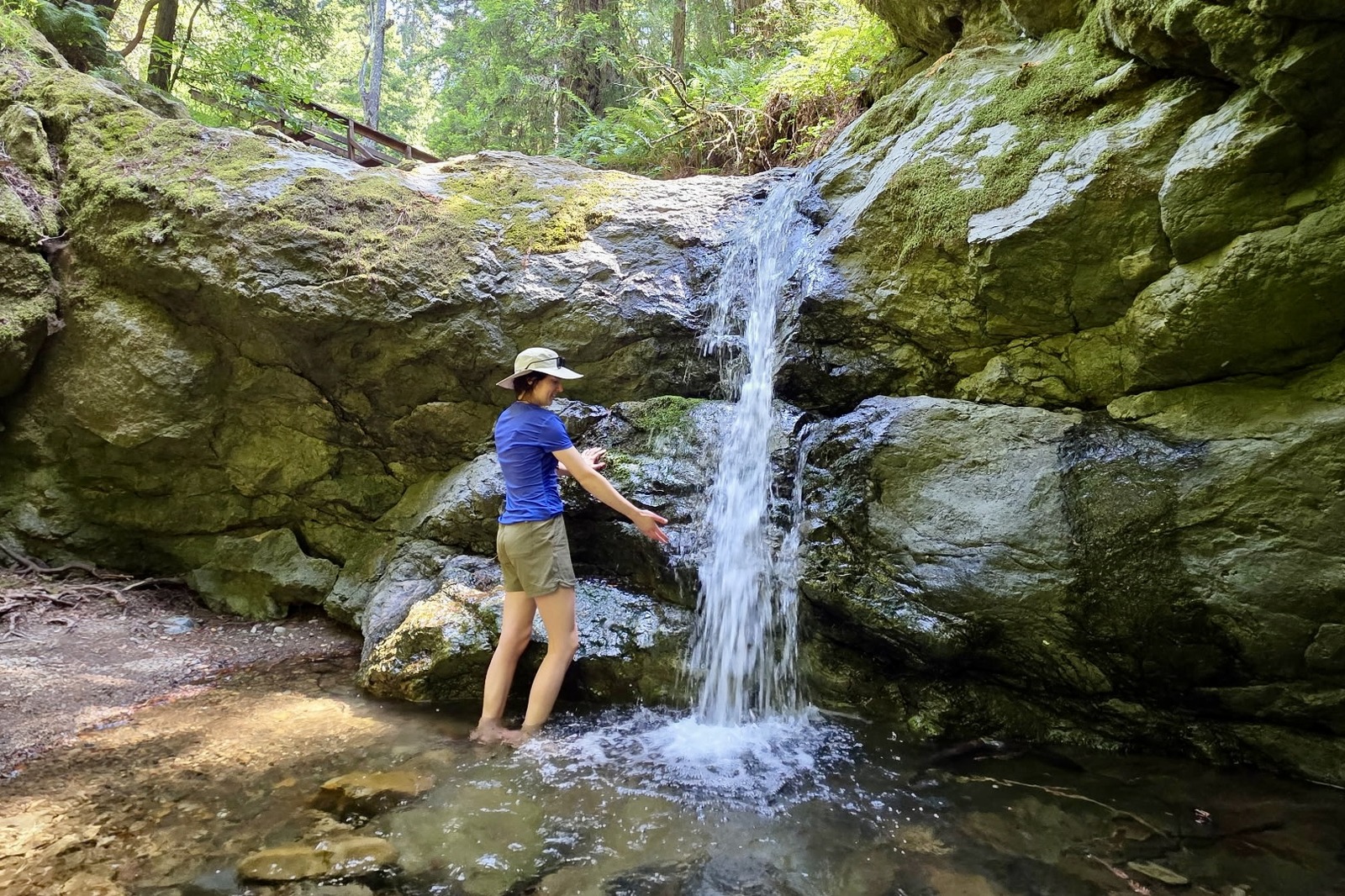 A woman stands next to a waterfall, wading in the pool below.