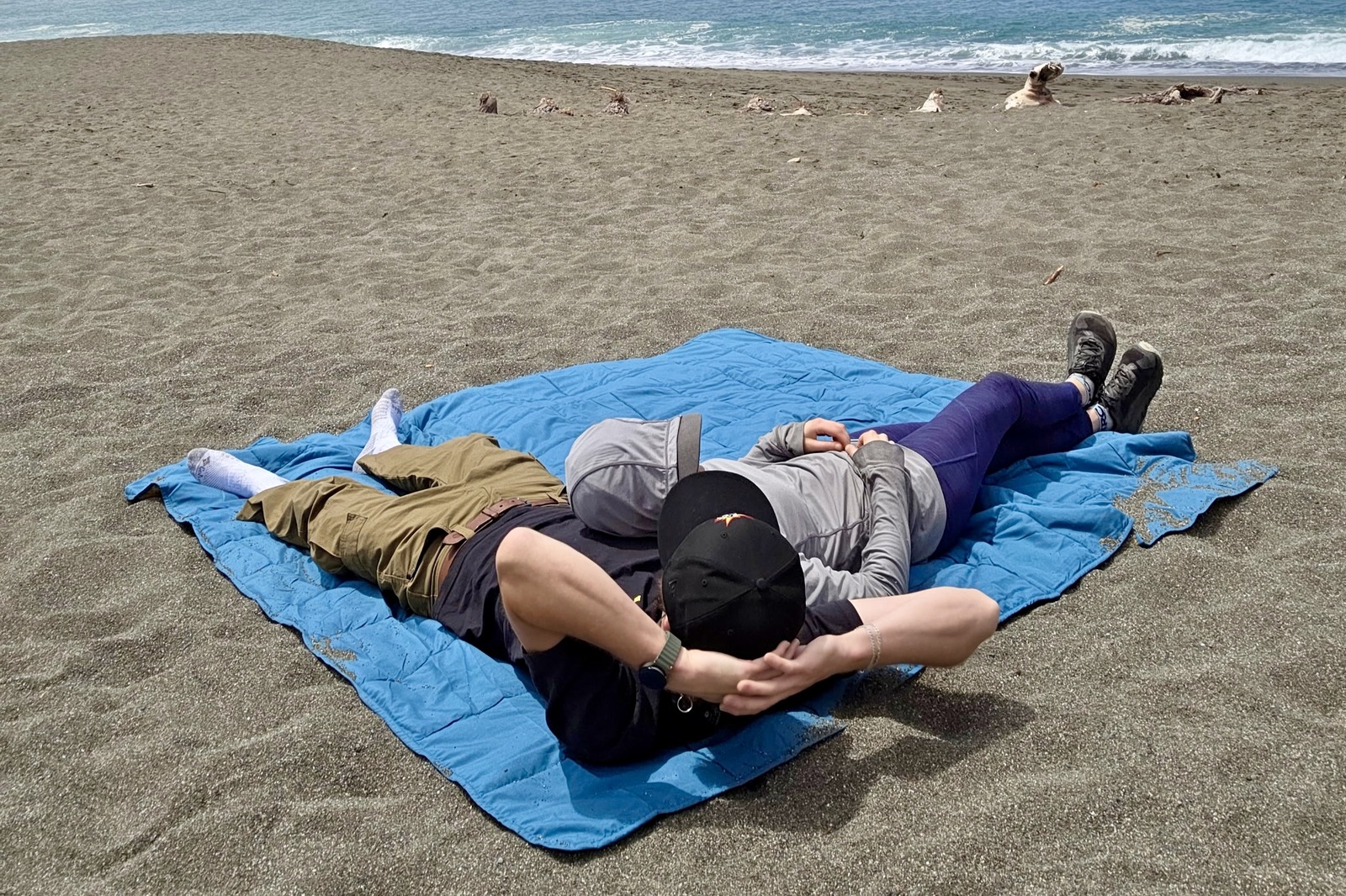 2 people lay on a blanket at the beach, one horizontally and one vertically, showing the size of the blanket