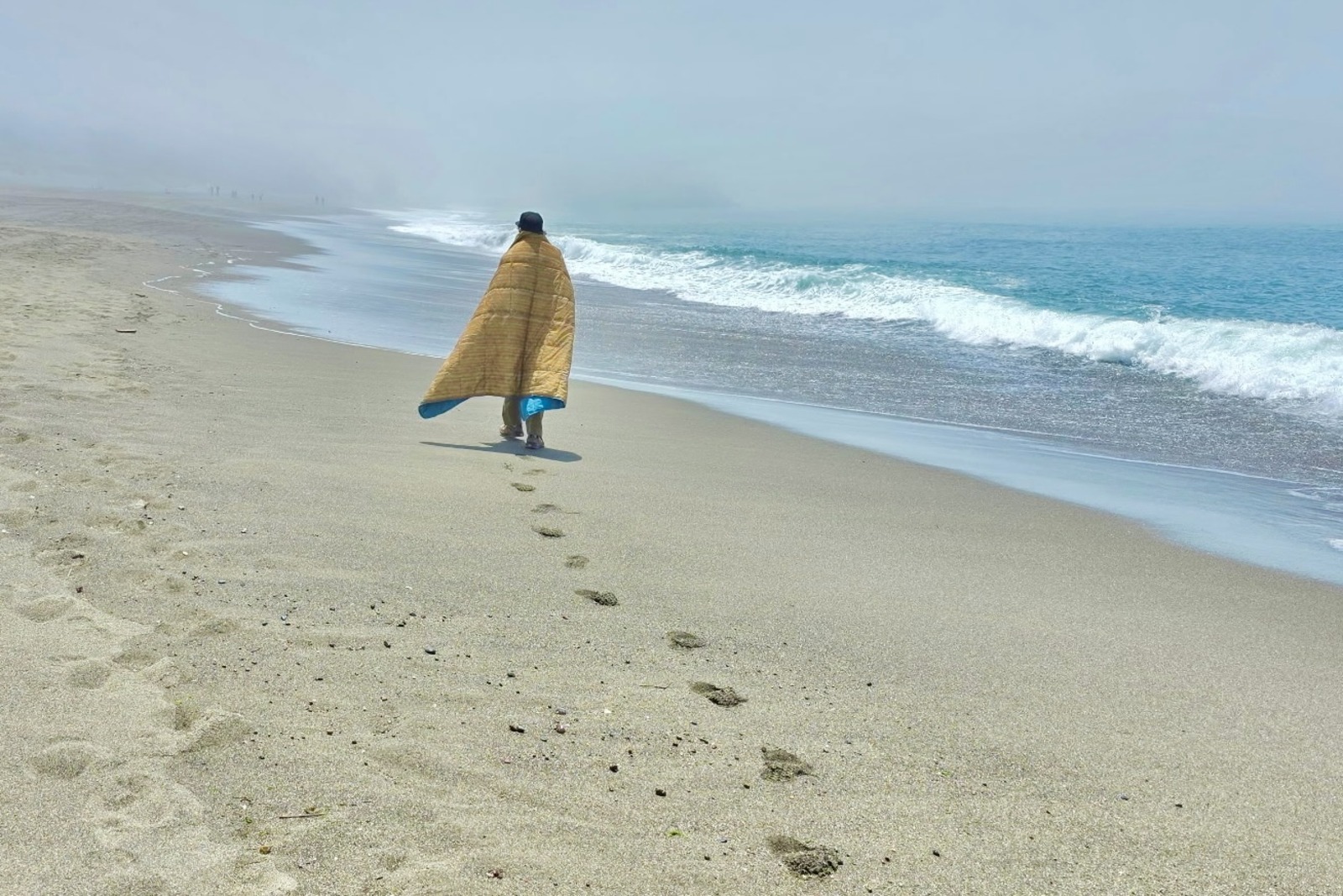 A man walks a long a foggy coastline wearing a blanket.