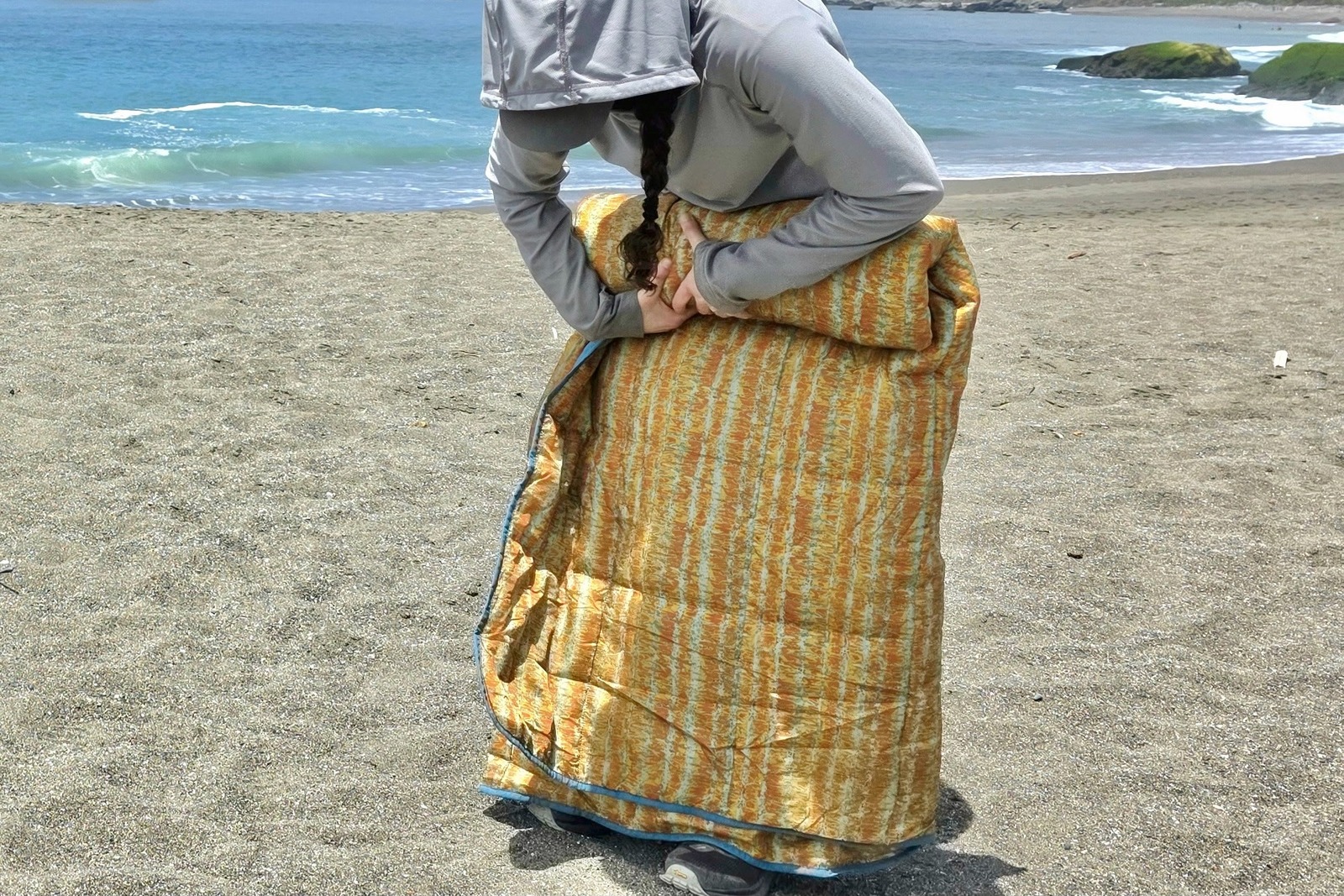 A woman rolls up her large camping blanket on the beach so she can pack it up.