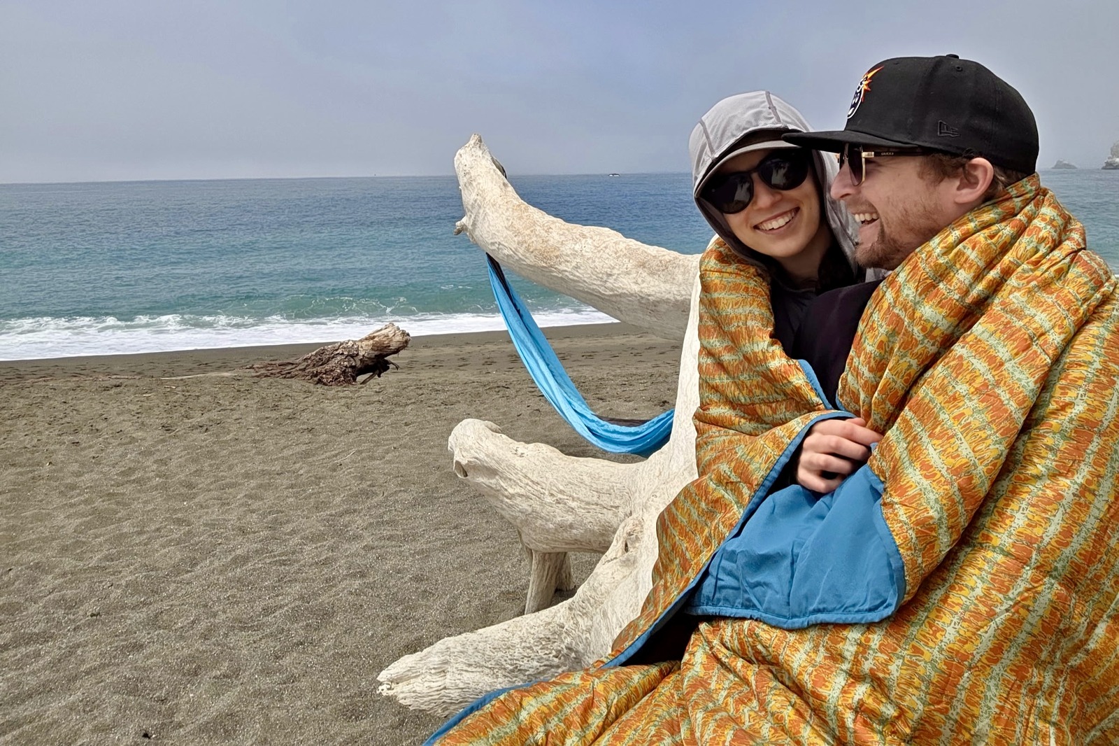 A man and woman sit on a log on the beach, wrapped up in a blanket.