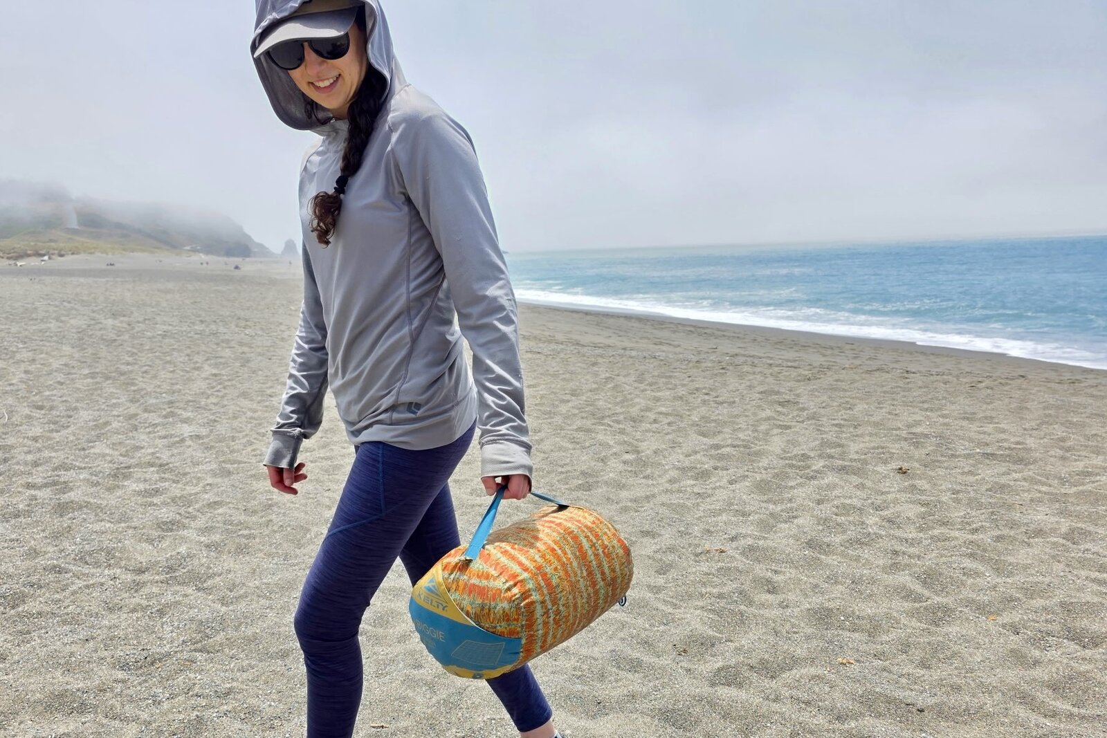 A woman carries her camping blanket across a beach, using the strap on the stuff sack.