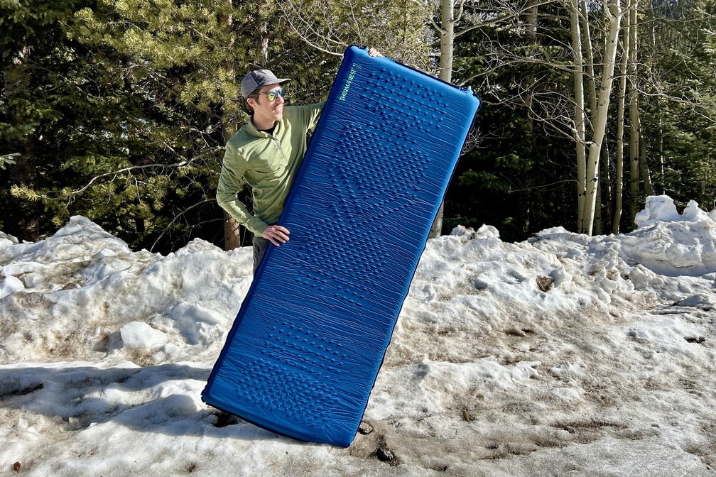 A man stands on a snowbank wearing sunglasses and a hat and holding up a camping mattress.
