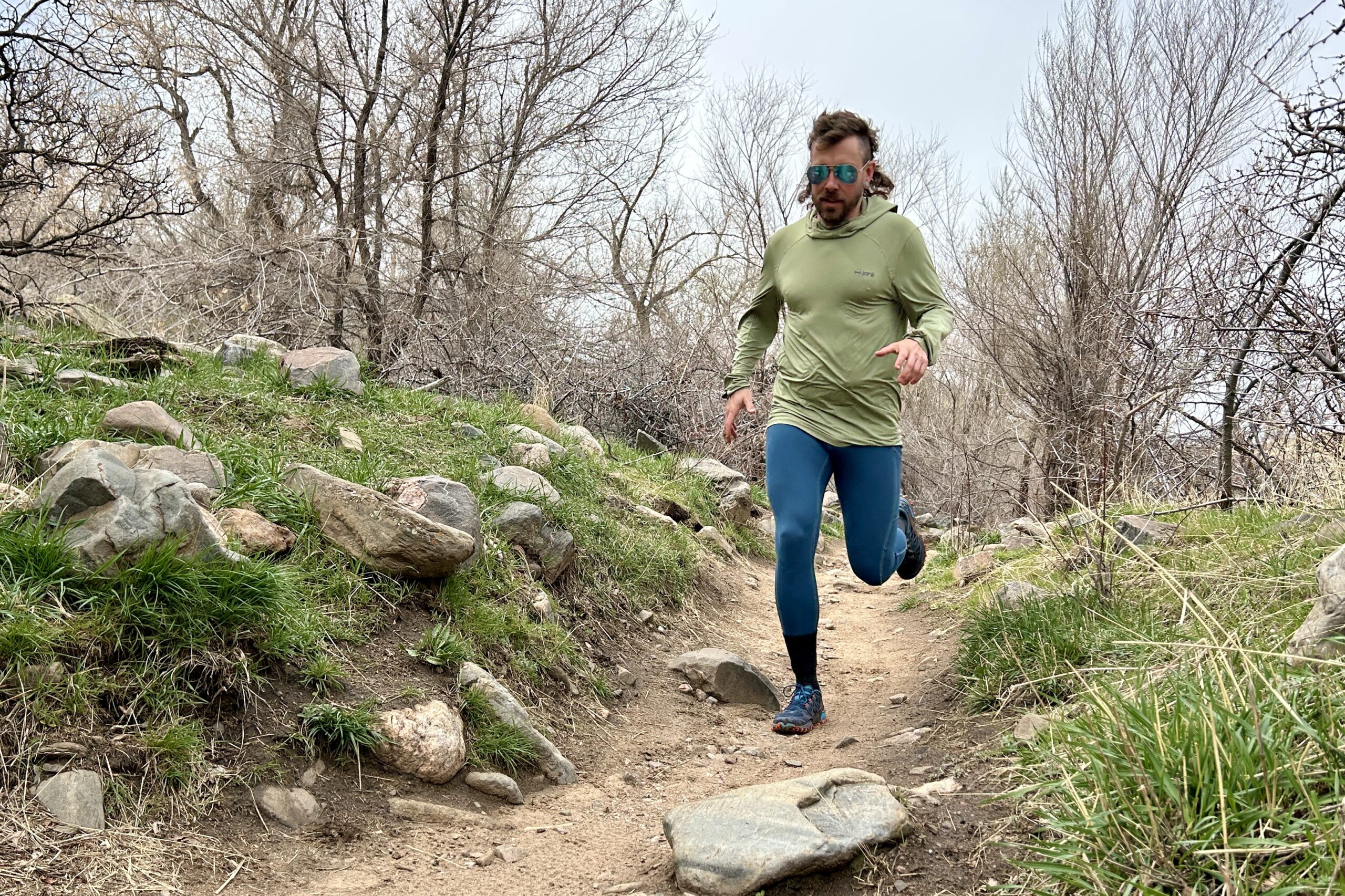 A man wearing a sun shirt, leggings, and aviator sunglasses runs down a trail in a wooded area.