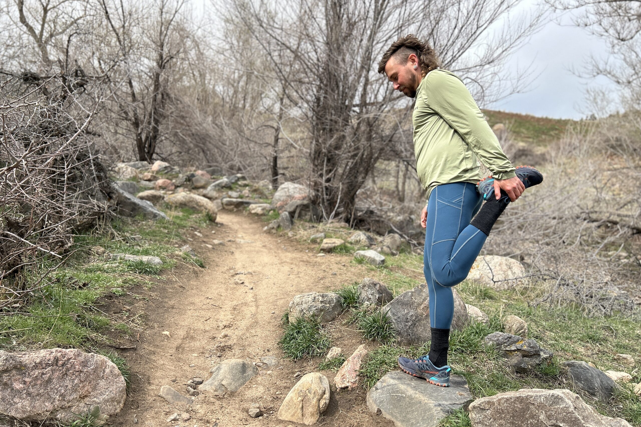 A man stretches one leg on a rock next to a trail in a wooded area.