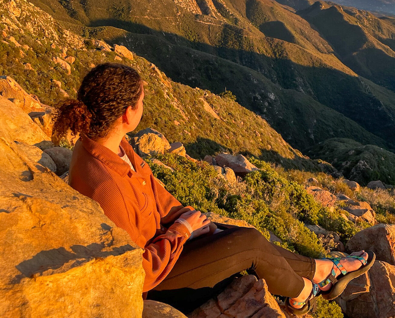 A woman sitting on a rocky outcropping in a green mountainous environment, wearing hiking clothes and sandals.