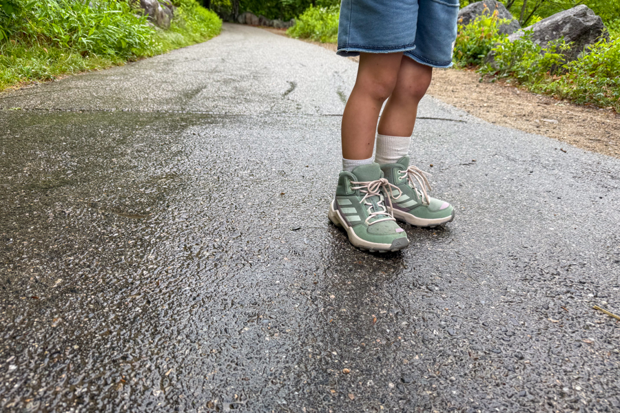 Child wearing Adidas Terrex AX4R Mid shoes standing on a wet asphalt trail after rain.