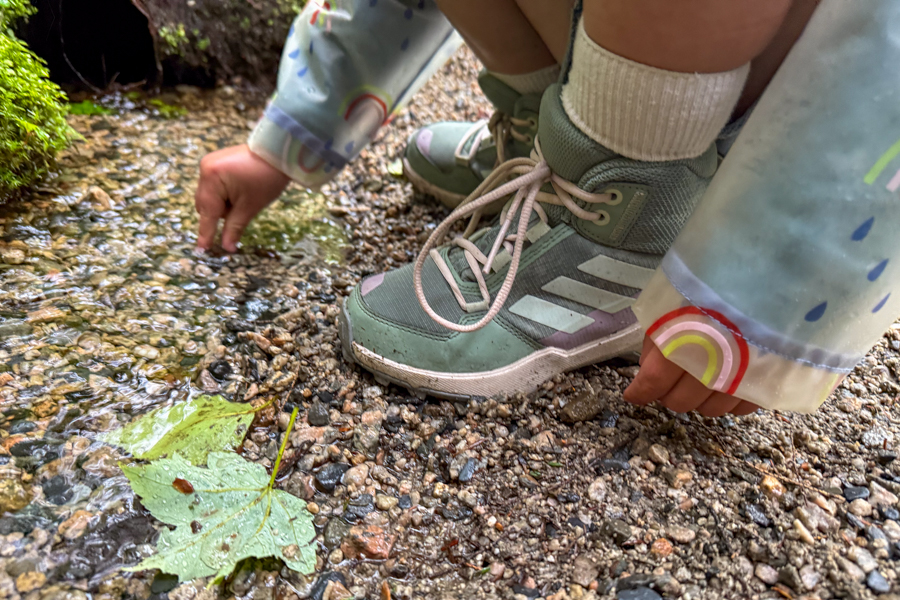 Child crouched at the edge of a shallow creek in Adidas Terrex AX4R Mid shoes and a colorful raincoat.