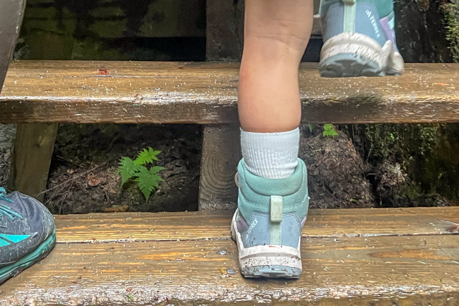 Child wearing Adidas Terrex AX4R Mid shoes climbing a wet wooden ladder on a forest trail.