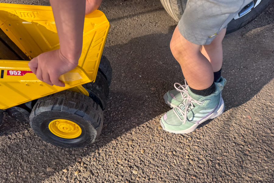 Child in Adidas Terrex AX4R Mid shoes playing with a large yellow toy dump truck on a dirt driveway.