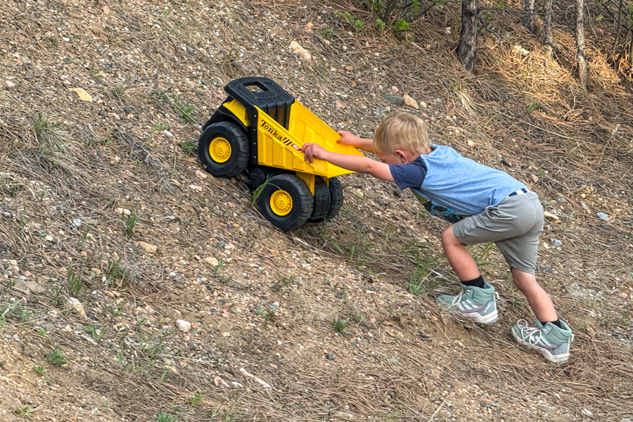 Child pushing a large toy dump truck up a steep, loose dirt trail while wearing Adidas Terrex AX4R Mid shoes.