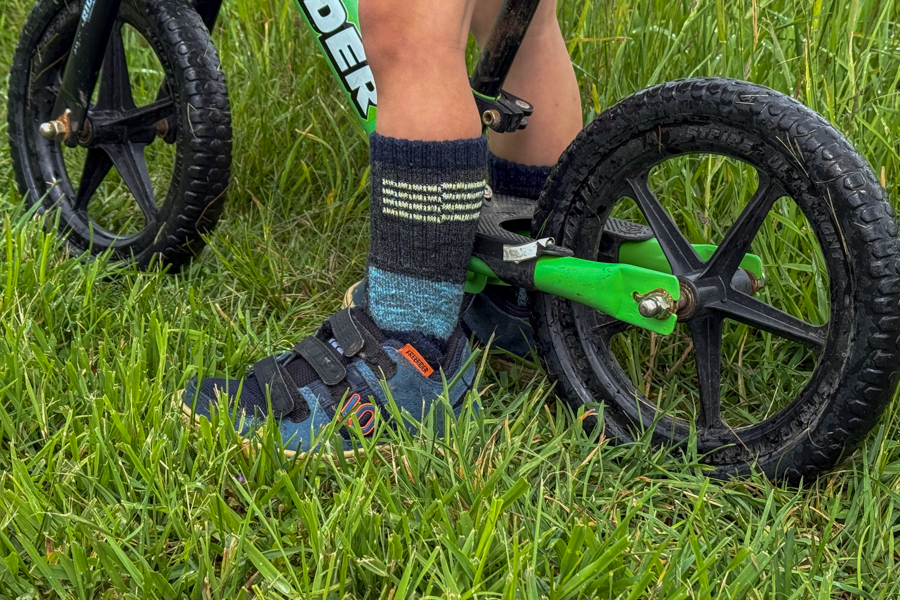 Close-up of child’s feet wearing Five Ten Freerider shoes while riding a green balance bike in the grass.