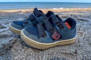 Close-up of worn Five Ten Freerider kids shoes sitting on sandy ground near a lake.