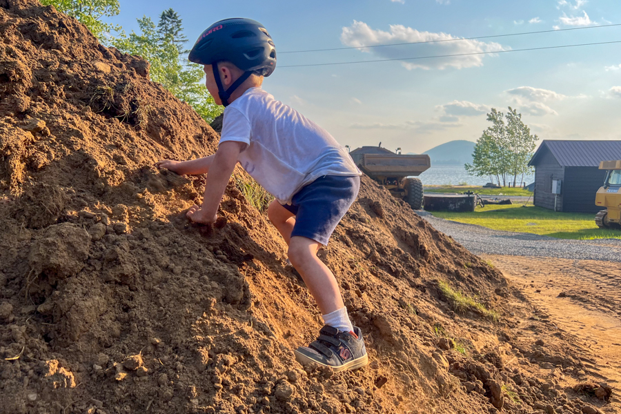 Child climbing a dirt hill in Freerider shoes with helmet on, construction equipment and lake in the background.