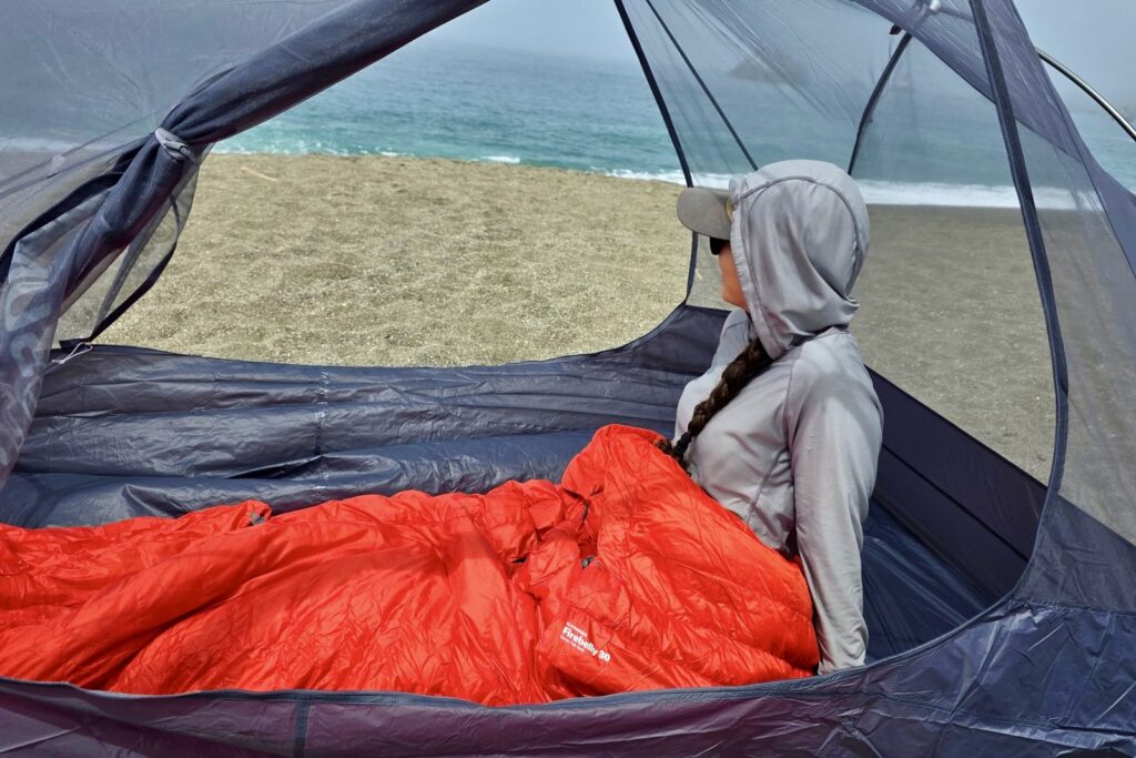 A woman sits in her tent on the beach, wrapped in her sleeping bag.