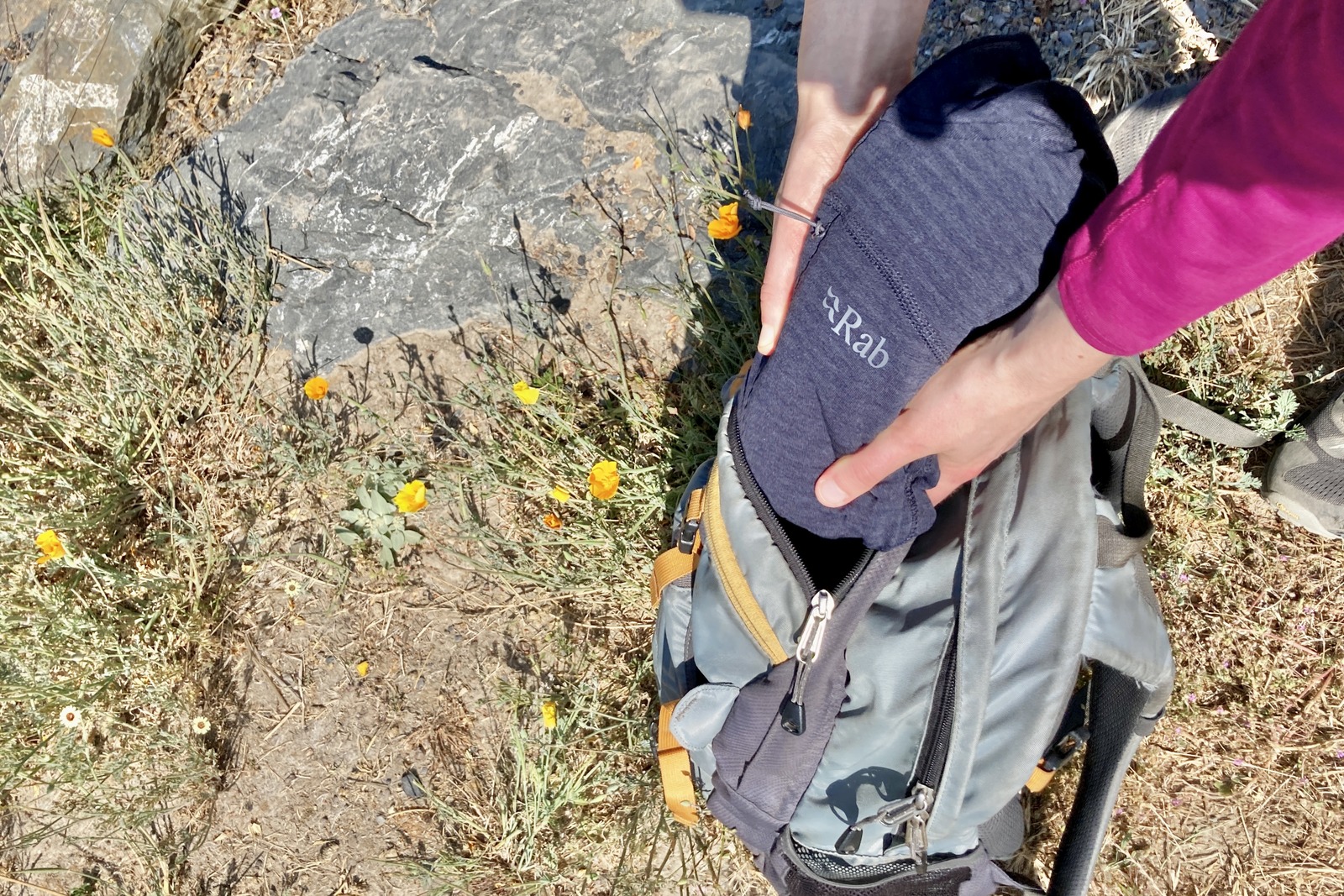 A woman packs up her fleece jacket into her daypack, next to some poppy flowers.