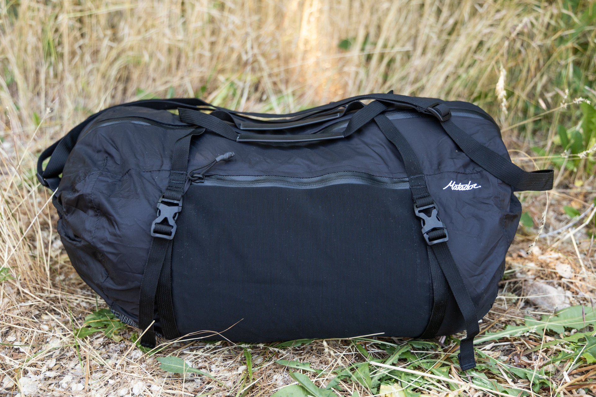 A side view of the Matador Freefly Packable Duffel. You can see two of the side compression straps and buckles, the mesh zipper pocket closed and empty, and the straps resting atop the bag. The matador logo is near the edge.