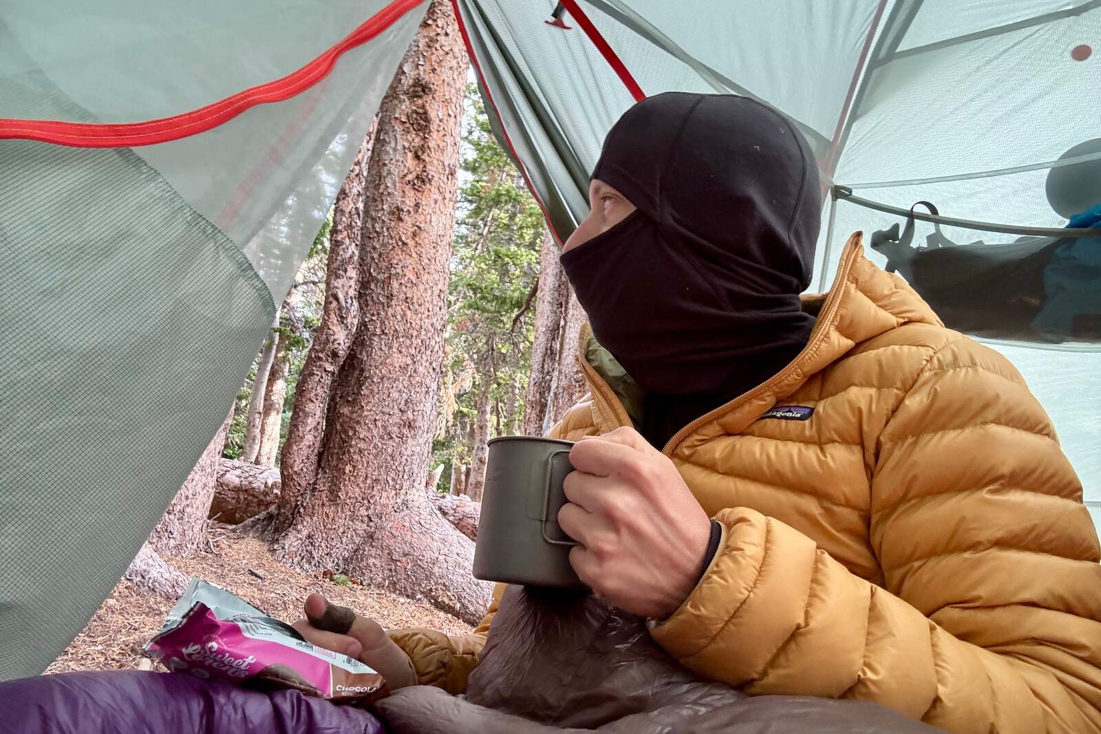 a hiker in a yellow puffy and a black balaclava hood sits in a tent looking out the door while sipping coffee